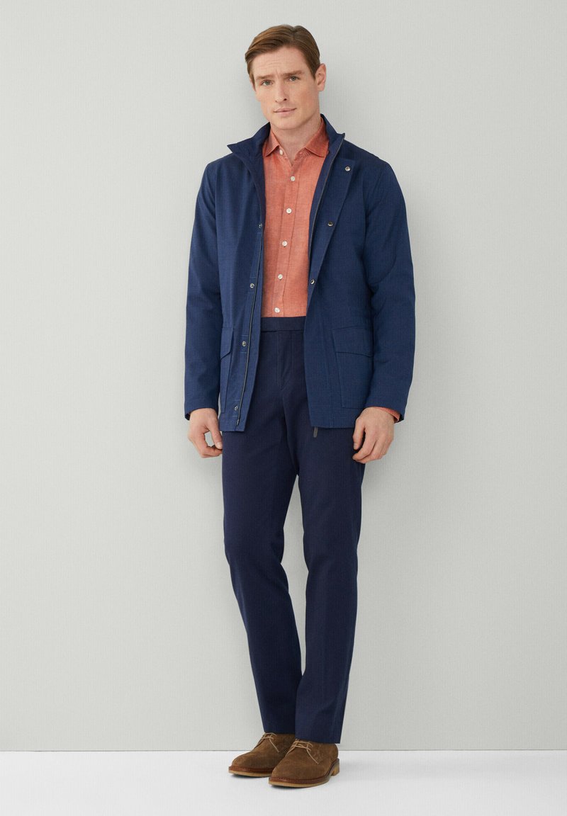 Veste bleu marine avec un col montant, portée sur une chemise corail à boutons et un pantalon bleu marine. Des chaussures en daim marron complètent la tenue.
