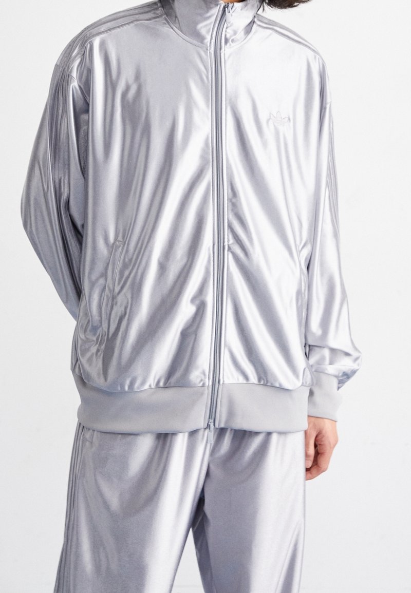 Osoba nosící lesklou stříbrnou sportovní bundu a kalhoty se žebrovanými manžetami a logem Adidas na hrudi na jednobarevném pozadí.