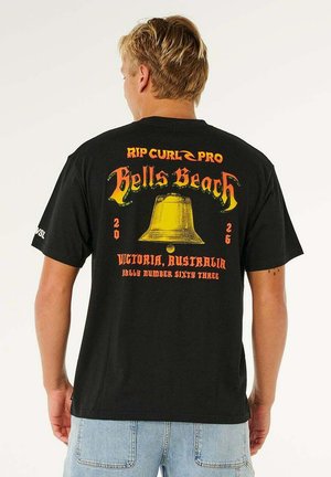 Persoon draagt een zwart Rip Curl Pro Bells Beach T-shirt met een gele belafbeelding en rode tekst, gecombineerd met lichtblauwe jeans.