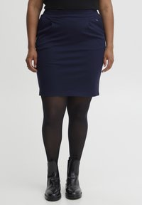 Jupe bleu marine mi-longue en tissu extensible avec poches latérales. Portée avec des collants opaques noirs et des bottines.