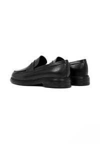Mocasines de cuero negro con un diseño elegante, suela plana de goma y sutiles acentos en la parte delantera y el collar. Textura suave, estilo minimalista.