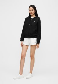 Svart hettegenser med glidelås foran, lomme og snorer, kombinert med hvite shorts med en logo på benet. Hvite joggesko fullfører antrekket.