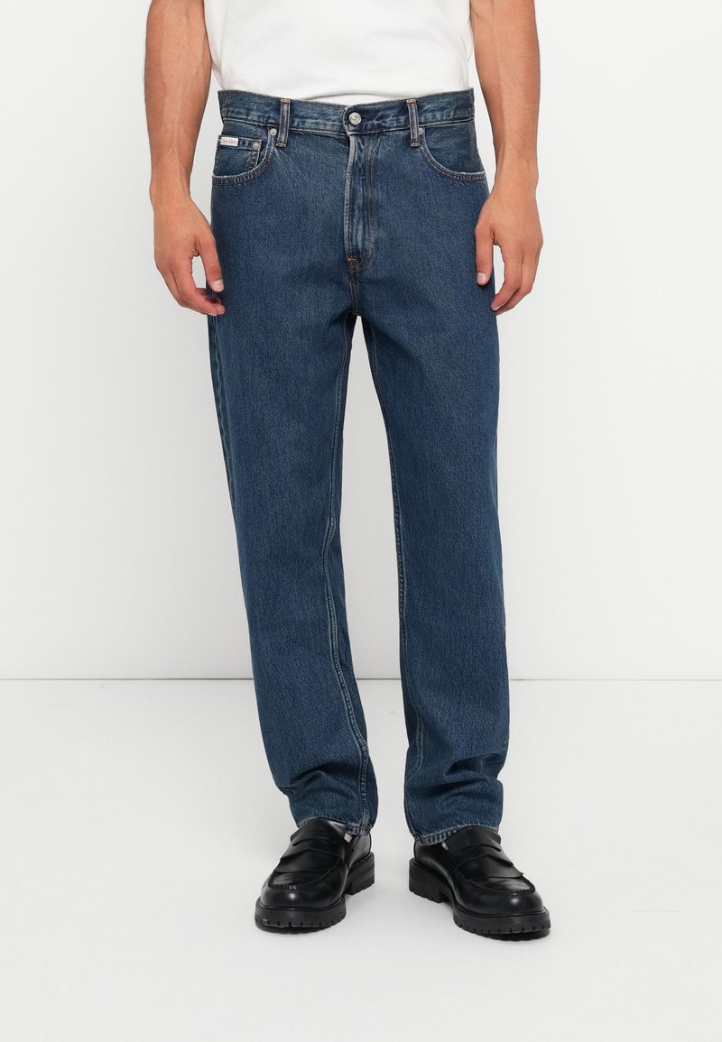Jean en denim bleu avec une coupe droite, cinq poches et une fermeture à bouton. Tissu texturé avec des détails de décoloration subtils.