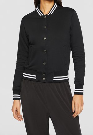 Femme portant une veste bomber noire boutonnée avec col, poignets et taille côtelés à rayures blanches, associée à un pantalon noir ample.