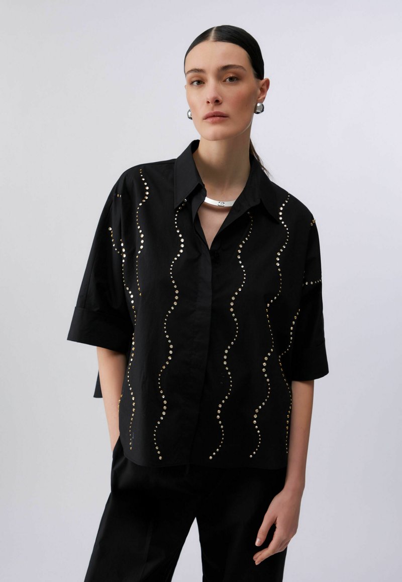 Femme aux cheveux noirs lisses portant une chemise noire à boutons ornée de clous argentés verticaux ondulés, un collier en argent, des boucles d'oreilles créoles et un pantalon noir.