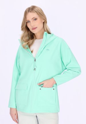 Blonde vrouw die een licht turquoise capuchonjas met zakken draagt, met één hand in een voorzak, tegen een effen witte achtergrond.