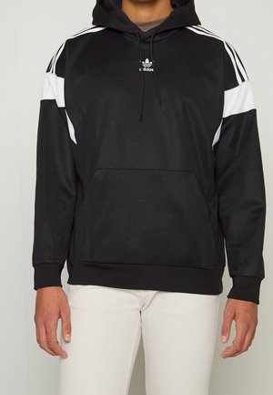 Svart Adidas-hoodie med vita ränder på axlar och armar, framficka, bärs med vita byxor på person från nacke till höfter.