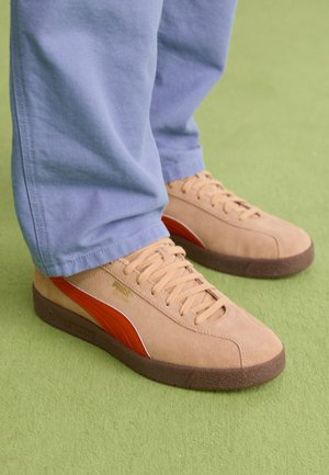 Baskets Puma en suède marron clair avec bande orange et semelle marron, portées avec un pantalon bleu clair sur un tapis vert.