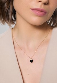 Złoty naszyjnik z łańcuszkiem, z czarnym serduszkiem w kształcie wisiorka, ozdobionym małym białym akcentem. Prezentowany na tle w odcieniu skóry.