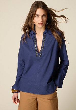 Pitkätukkainen ruskeahiuksinen nainen pukee tummansinisen helmirivillä koristellun V-pääntiepaidon ja vaaleat housut, seisoo neutraalin taustan edessä.