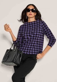 Vrouw met grote zwarte zonnebril, paars en zwart houndstooth sweater, zwarte broek, en een zwarte leren handtas met metalen studs.
