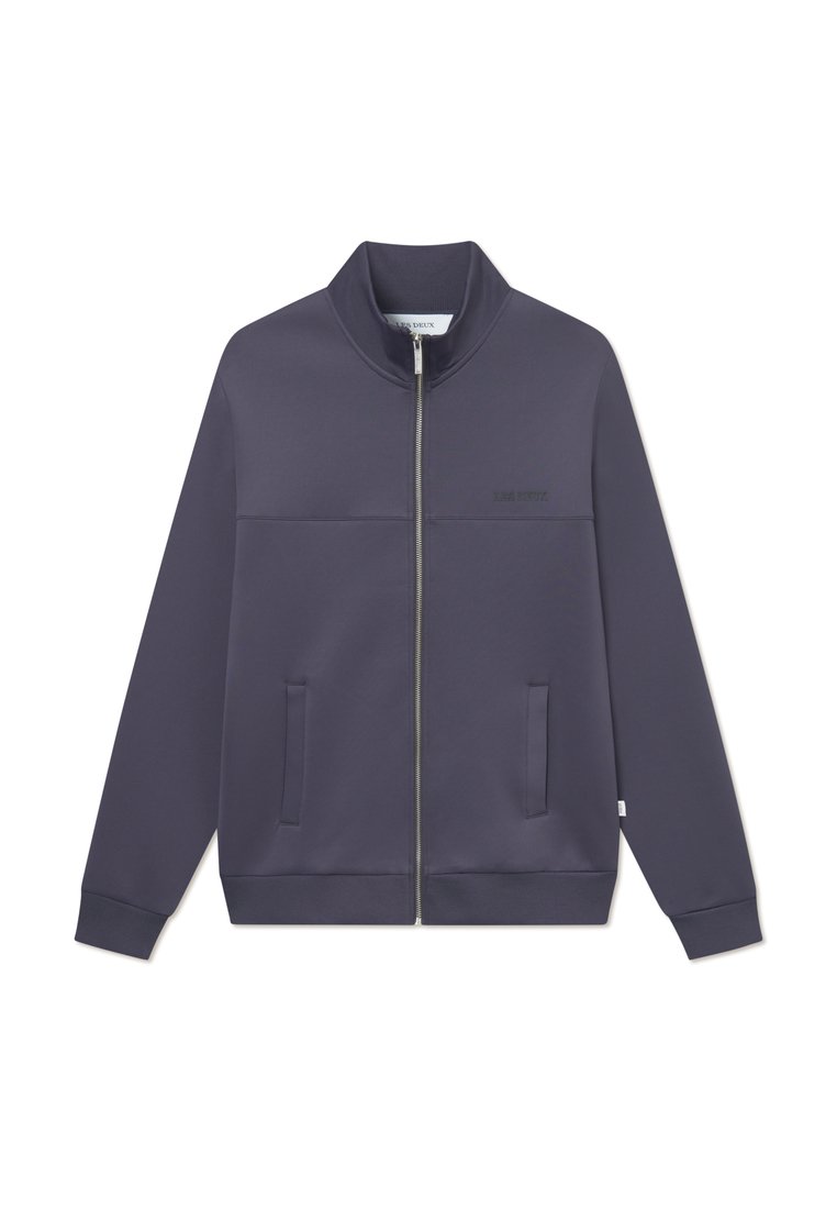 Chaqueta de color púrpura oscuro con cremallera, cuello alto, bolsillos laterales, puños acanalados y un discreto detalle de logo en el pecho. Textura de tela suave.