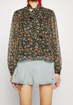 Blouse noire transparente à motif floral avec manches longues et nœud au col, associée à un short en jean bleu clair plissé.