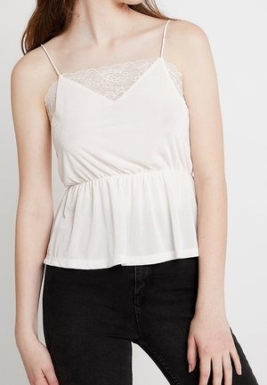 Vrouw draagt een wit mouwloos topje met kanten afwerking en dunne bandjes en een zwarte broek, staand tegen een effen achtergrond.