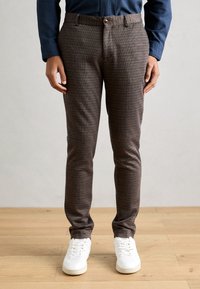 Brown patterned trousers featuring a slim fit, textured fabric, belt loops, and a front button closure, paired with white sneakers.