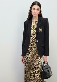 Čierne sako s zlatým gombíkom a logom, skombinované s leopardím šatami. Modelka drží čiernu textúrovanú kabelku.