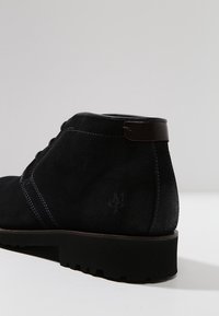 Stivali alla caviglia in suede neri con punta tondeggiante, accentuati da un dettaglio in pelle marrone sul tacco e cuciture blu a contrasto. Dettaglio della superficie texture visibile.