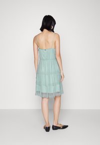 Trojvrstvá mátová šaty s tenkými ramínky, krajkovým lemem a upnutým korzetem, se zapínáním na knoflíky na zadní straně. Černé ploché boty doplňují outfit.