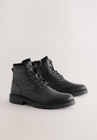 Schwarze Leder-Stiefeletten mit glatter Oberfläche, ausgestattet mit Schnürsenkeln, einem seitlichen Reißverschluss und einer profilierten Gummisohle. Kontrastierender textiler Besatz am oberen Rand.