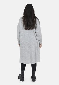 Lange grijze gebreide cardigan met ribbelstructuur, open rug, verlaagde schouders en een rechte snit. Gedragen met zwarte leggings en chunky laarzen.