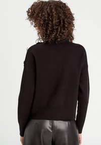 Pull en tricot noir avec une coupe décontractée, un ourlet côtelé et des zips sur les côtés. Montre la texture et les détails vue de dos.
