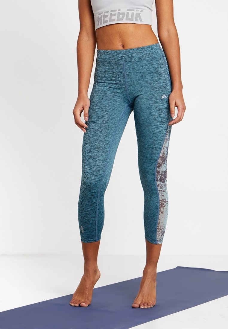 Kvinna som står barfota på en yogamatta, klädd i blå mönstrade leggings och en vit kortärmad Reebok-top, mot en plain vit bakgrund.