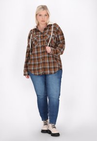 Brązowa i szara koszula w kratę z dwiema przednimi kieszeniami, długimi rękawami i kremowym kapturem, w połączeniu z niebieskimi dżinsami i beżowymi butami na sznurówki.