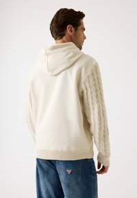 Sudadera crema con mangas de punto trenzado texturizado. Cuenta con capucha con cordones y dobladillo acanalado. Combinada con jeans de mezclilla azules.