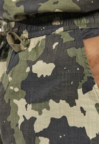 Close-up of hand in pocket of green, black, and beige camouflage patterned pants with drawstring waist.