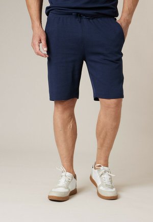 Homme portant un short bleu marine et des baskets blanches, debout les mains dans les poches devant un fond uni.