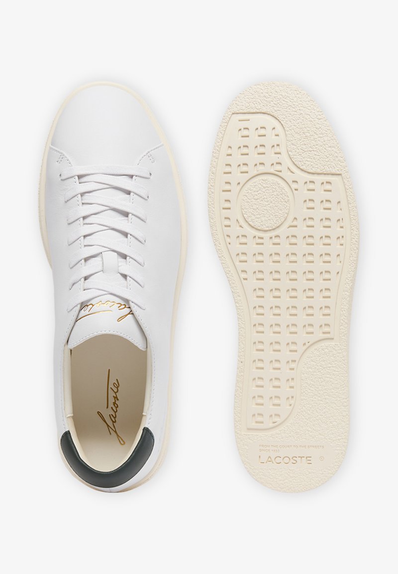 Sneaker in pelle bianca con inserto sul tallone nero, logo in corsivo dorato all'interno e suola crema testurizzata con marchio Lacoste visibile.