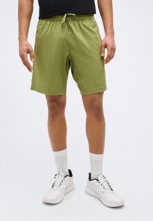 Mees, kes kannab oliivrohelisi paelaäärega lühikesi pükse, valgeid sokke ja valgeid tossusid, seisab halli ühevärvilise tausta ees.