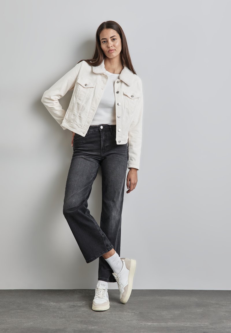 Vit cordjacka i cordflät med knäppningar, parat med svarta vida jeans och vita sneakers. Släta texturer och avslappnad passform.