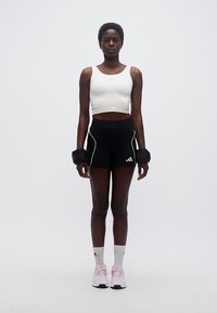 Γυναίκα όρθια, φορώντας λευκό crop top, μαύρο αθλητικό σορτς, λευκές κάλτσες, ροζ αθλητικά παπούτσια και μαύρα βαράκια καρπού, με φόντο απλό λευκό.