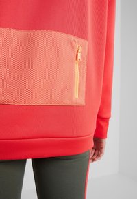 Sudadera deportiva roja con un bolsillo de malla texturizada y una cremallera diagonal, junto con un dobladillo acanalado. El diseño incorpora colores contrastantes.