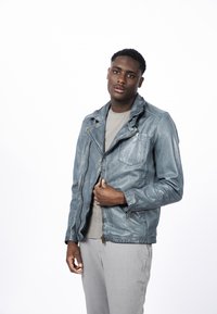 Chaqueta de cuero azul con cremallera frontal, hombreras y dos bolsillos con cremallera. Estilizada con una camisa gris clara y pantalones grises.