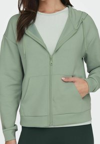 Žena v khaki zelené mikině s zipem, předními kapsami, kapucí a šňůrkami, kterou má oblečenou na světle zeleném tričku.