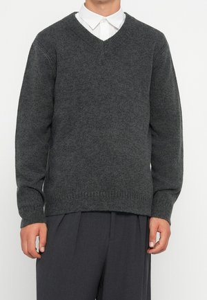 Man draagt een donkergrijze V-hals trui over een wit overhemd met kraag en donkergrijze broek tegen een effen achtergrond.
