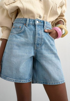 Personne portant un short en jean bleu clair avec un bouton et une fermeture éclair, un haut beige à manches longues et des bracelets colorés à un poignet.