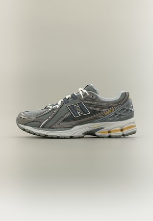 Zapatilla de deporte gris y negra con cordones blancos, suela gruesa acolchada y logo de la marca en el lateral, colocada sobre un fondo liso beige.