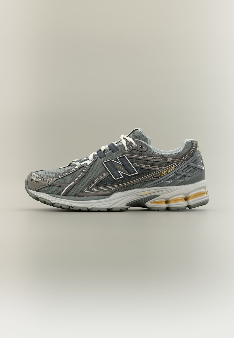 Γκρι και μαύρο αθλητικό παπούτσι με λευκά κορδόνια, παχύ απορροφητικό πέλμα και λογότυπο της μάρκας στο πλαϊνό μέρος, τοποθετημένο σε απλό μπεζ φόντο.