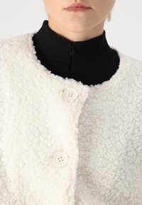 Veste en polaire blanche texturée avec un col rond, fermeture à boutons et un haut en col côtelé noir en dessous.