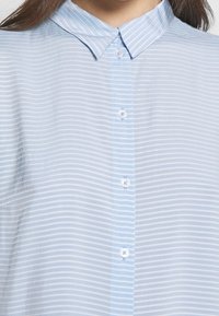 Camicia azzurro chiaro con bottoni, realizzata in tessuto sottile con strisce orizzontali bianche, dotata di un colletto a punta e bottoni bianchi.