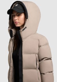 Chaqueta tipo puffer beige con una gran capucha, cierre de cremallera y detalles en negro. Presenta un tejido acolchado y puños elásticos para mayor calidez.
