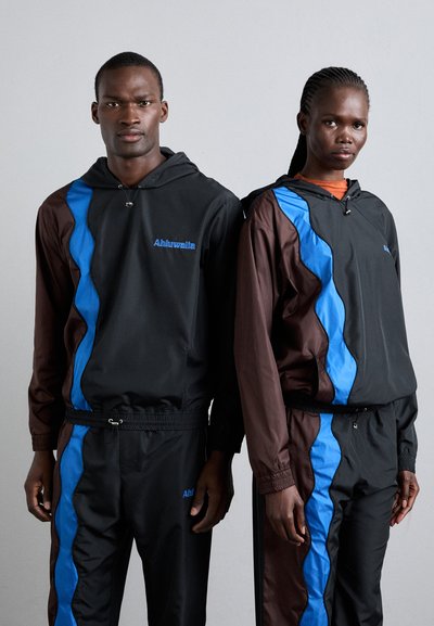Sorte og brune hættetrøje tracksuits har blå sidelister og snoretræk i livet. Logoet "Ahluwalia" er synligt på begge sæt.