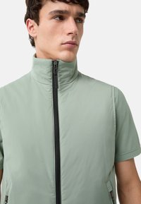Hellgrüne ärmellose Jacke mit hochgeschlossenem Kragen, Reißverschluss vorne und seitlichen Taschen. Glattes Material mit matter Oberfläche.