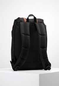 Sac à dos en tissu noir avec des bretelles rembourrées et une poignée supérieure. Présente une texture lisse et un design minimaliste.