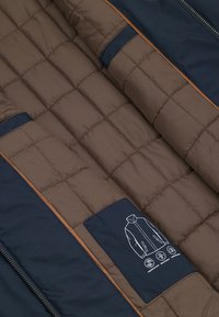 Brązowa pikowana tkanina z granatowymi akcentami, zawierająca etykietę z ilustracją projektu kurtki i symbolami pielęgnacji. Teksturowana powierzchnia z liniowymi wzorami.