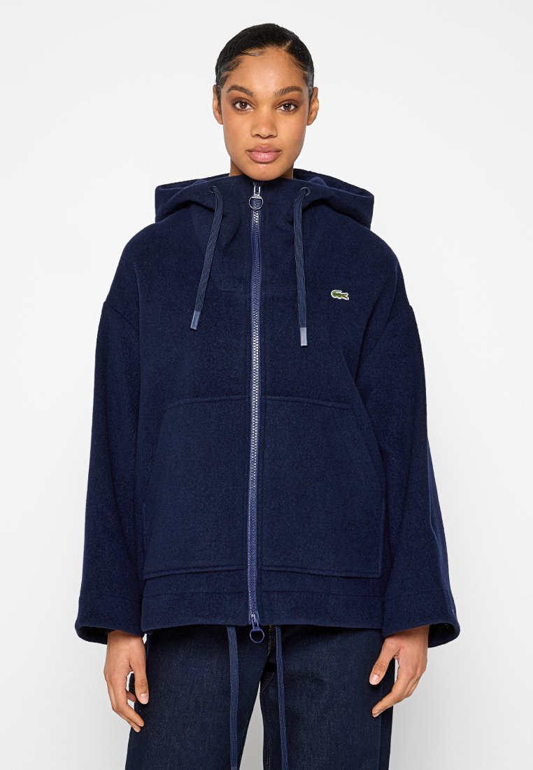 Marineblaue Kapuzenjacke mit Reißverschluss aus weichem Material, ausgestattet mit einer großen Fronttasche und einem kleinen Lacoste-Logo auf der linken Brust.