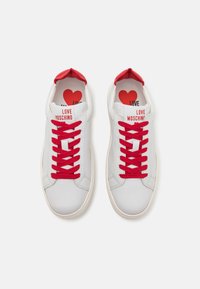 Fehér sportcipők piros fűzőkkel és díszítésekkel. Jellemzője a lekerekített orr, sima bőr, valamint a "LOVE MOSCHINO" felirat a nyelvén.